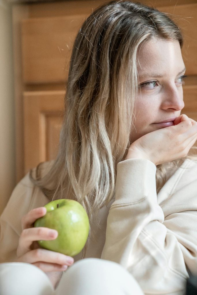 A young woman sits indoors, holding a green apple and gazing thoughtfully. Soft lighting and neutral tones.