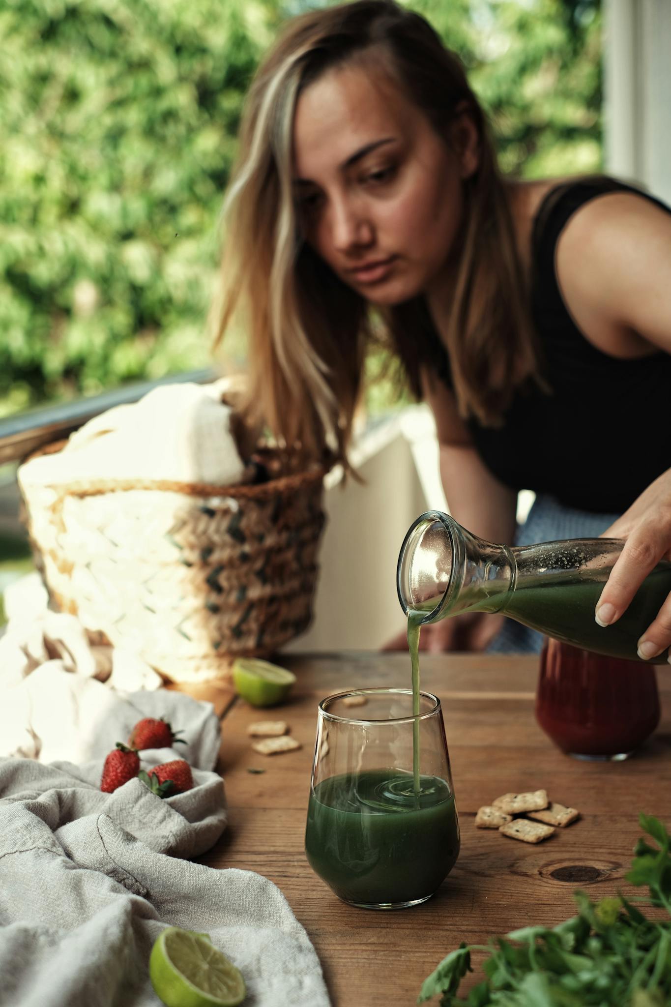 Young woman pours fresh green juice into a glass on a sunny outdoor patio setting.